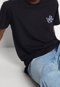 Czarna bawełniana koszulka z okrągłym dekoltem, z białym graficznym nadrukiem na piersi, który brzmi "ON VACATION CLUB". Luźny krój.