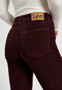 Burgundy corduroy jeans with a textured surface, featuring a leather patch with "Lee" logo at the waist and back pockets with stitching details.