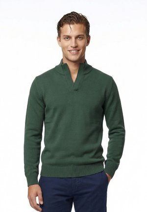 Ung mand smilende, iført en mørkegrøn strikket sweater med V-udskæring og marineblå bukser, stående foran en hvid baggrund.