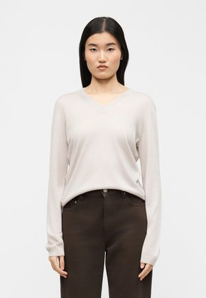 Mladá žena s rovnými čiernymi vlasmi, ktorá má na sebe svetlo béžový sveter s výstrihom do V a tmavohnedé nohavice, stojaca pred jednoduchým pozadím.