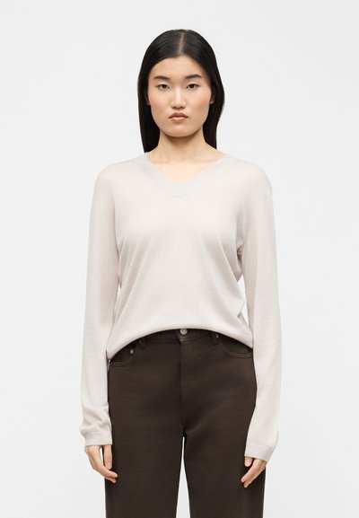Jeune femme aux cheveux noirs raides portant un pull col en V beige clair et un pantalon marron foncé, debout devant un fond uni.
