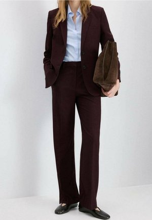 Vrouw in donker bordeauxrood pak met lichtblauw overhemd, met een grote bruine suède clutch, zwarte platte schoenen, staand tegen een witte muur.