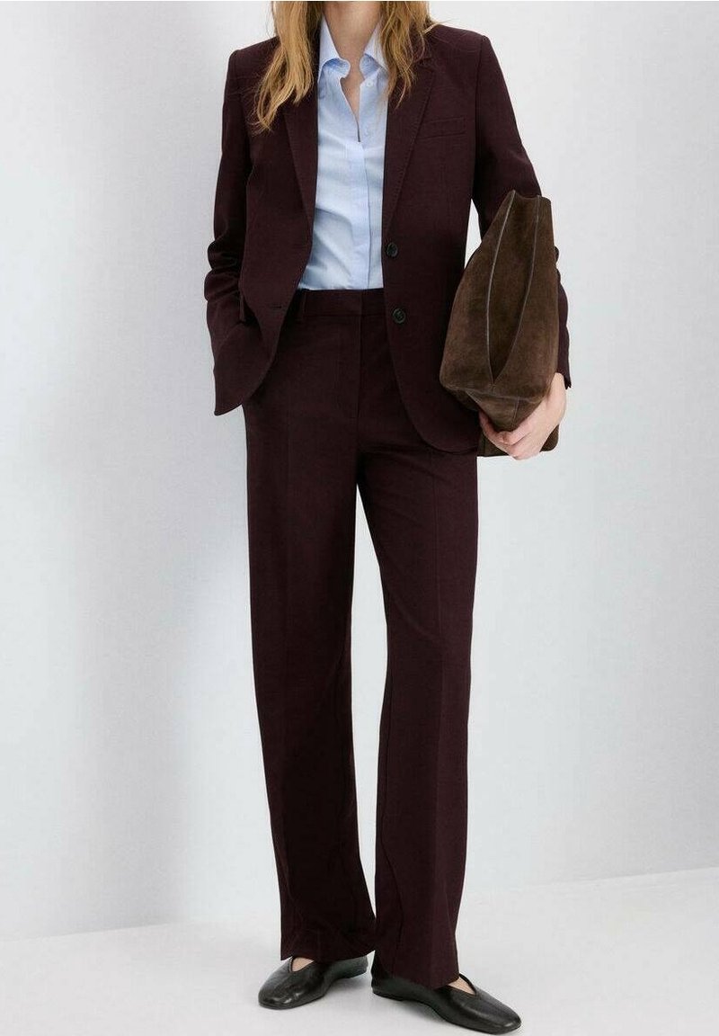 Femme en costume bordeaux foncé avec une chemise bleu clair, tenant une grande pochette en suède marron, portant des chaussures plates noires, debout contre un mur blanc.