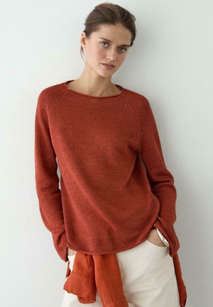 Junge Frau mit Händen in den Taschen, die einen rostfarbenen Strickpullover und hellbeige Hosen vor einem schlichten hellen Hintergrund trägt.