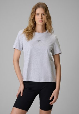 Kobieta o falujących włosach, ubrana w jasnoszary t-shirt i czarne szorty kolarskie, stojąca na tle jednolitego szarego tła.