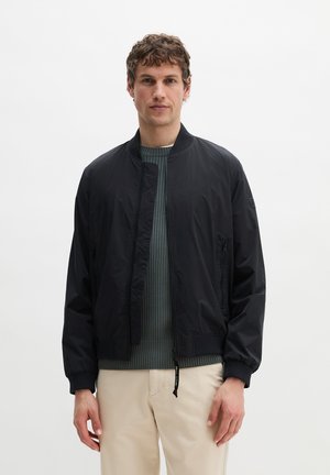 Junger Mann trägt eine schwarze Bomberjacke über einem grauen, gerippten Pullover und beigen Hosen, steht vor einem schlichten weißen Hintergrund.