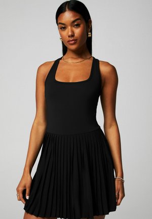 Frau mit langen schwarzen Haaren, die goldene Creolen, eine Kettenhalskette und ein ärmelloses schwarzes Plissee-Kleid trägt, steht vor einem grauen Hintergrund.