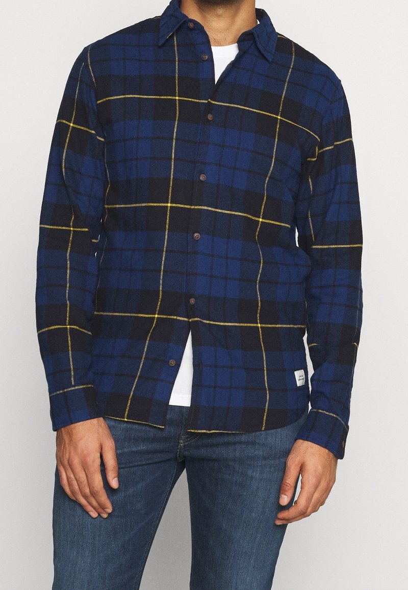 Homme portant une chemise à carreaux bleue, noire et jaune sur un t-shirt blanc et un jean bleu, se tenant avec les mains détendues sur les côtés.