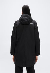 Μαύρο αδιάβ ροχο μπουφάν με κουκούλα, με ορατό λογότυπο στην πλάτη, μακριά μανίκια και ίσια γραμμή που φτάνει κάτω από τη μέση.