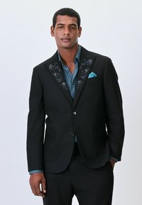 Schwarzer Blazer mit floraler Stickerei an den Revers, kombiniert mit einem blauen Button-up-Hemd. Verfügt über ein Einstecktuch und zwei Vorderknöpfe.