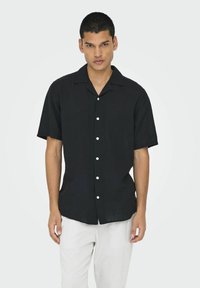 Camisa negra de manga corta en corte relajado, con cuello abierto y botones blancos al frente. Textura suave con un ligero brillo.