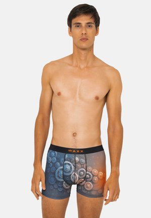 Jeune homme debout, portant un boxer avec un motif d'engrenages bleu et orange, ceinture noire avec l'inscription "WAXX".