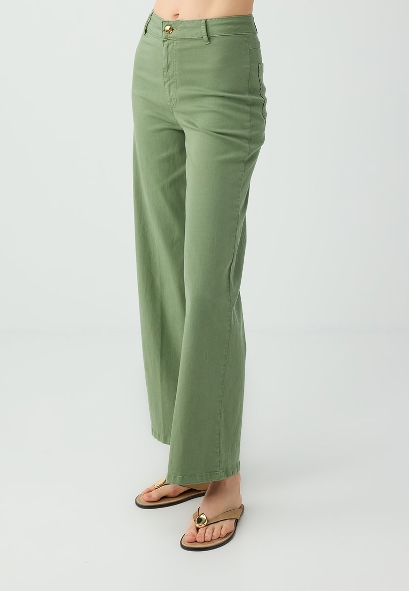 Femme portant un pantalon large taille haute vert olive et des sandales beiges avec des décorations circulaires dorées sur fond blanc.