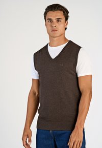 Brauner ärmelloser V-Ausschnitt-Pullover aus weichem Material, getragen über einem weißen T-Shirt. Mit geripptem Ausschnitt und Saum, der einen lässigen Schnitt unterstreicht.