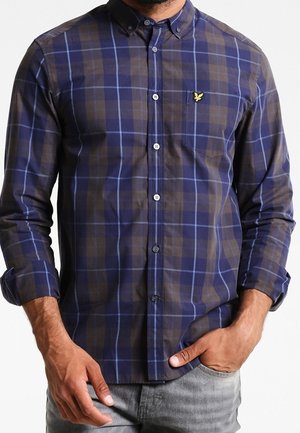 Homme portant une chemise à carreaux marron et bleue à boutons avec les manches retroussées et un pantalon gris, main dans la poche.