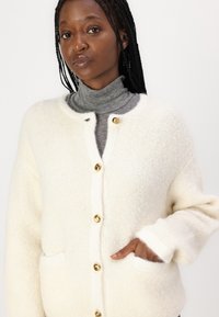 Cardigan boutonné en crème, fabriqué dans un tissu doux et texturé. Dispose de boutons dorés et de deux poches latérales.
