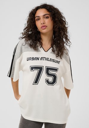 Jeune femme aux cheveux foncés et bouclés portant un t-shirt sportif blanc et gris avec "Urban Athleisure" et le numéro 75 imprimés sur le devant.