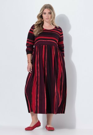 Lang gestreiftes Kleid in Rot und Schwarz mit eng anliegender Oberseite und schwingendem Rock. Verfügt über lange Ärmel und einen runden Halsausschnitt. Kombiniert mit roten Ballerinas.