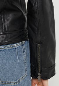 Veste en cuir noir avec une texture lisse, dotée d'un poignet zippé et d'un jean en denim bleu contrastant avec des coutures jaunes.