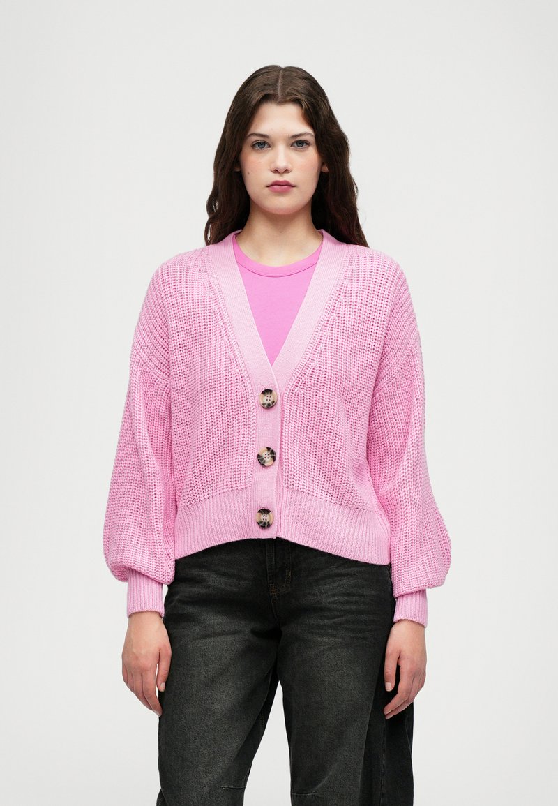 Lyserosa strikket cardigan med dyp V-hals, tre store knapper, ballongsleeves og cropped hem. Brukt over en rosa topp og svarte bukser.