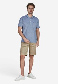 Helles blaues Polohemd mit kurzen Ärmeln, ausgestattet mit einer Brusttasche; kombiniert mit beigen Shorts und weißen Sneakern, stehend vor einem schlichten Hintergrund.