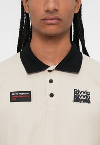 Beige polo shirt with a black collar, featuring a black logo and a patch that reads "Rivvia Projects - Active Discovery," with three buttons.