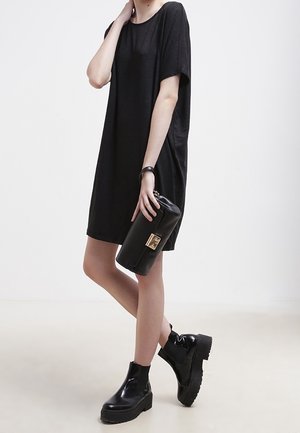 Zwart oversized t-shirt jurk met korte mouwen, zachte textuur. Model houdt een zwarte clutch met gouden hardware vast, gecombineerd met chunky zwarte laarzen.