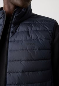 Colete acolchoado azul marinho com colarinho alto, apresentando costura acolchoada horizontal e um logotipo subtil do lado esquerdo. Detalhe do fecho visível.