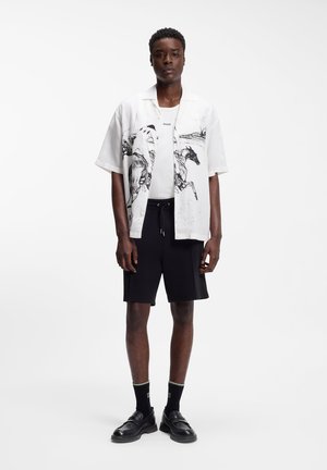 Mann trägt weißes Kurzarmhemd mit schwarzem Pferdeaufdruck, weißes T-Shirt, schwarze Shorts, schwarze Socken und schwarze Schuhe, steht vor weißem Hintergrund.