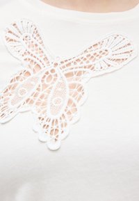 Λευκό ύφασμα με λεπτομερή εφαρμογή δαντελένιου μοτίβου πεταλούδας κοντά στο ντεκολτέ που δείχνει το περίτεχνο ανοιχτό σχέδιό του.