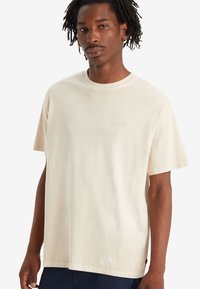 T-shirt en coton couleur crème avec une coupe décontractée, des manches courtes et un logo brodé subtil sur la poitrine. Texture douce, design casual.