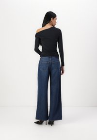 Haut noir à manches longues avec un design d'épaule asymétrique, associé à un jean bleu à jambes larges. Le modèle porte des escarpins noirs.
