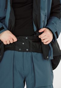 Blaues Sakko mit schwarzem Futter und Druckknopfverschluss, getragen über einem schwarzen Hemd. Darunter blaue Hose mit gefaltetem Design und seitlichen Reißverschlüssen.