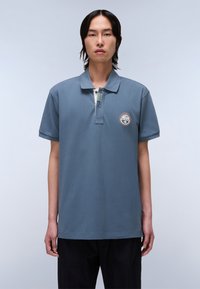 Šedé bavlněné polo tričko s límečkem, krátkými rukávy a vyšitým odznakem na levém prsu. Disponuje zapínáním na dvě knoflíky.