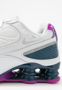 Bílé sportovní boty s vrškem z prodyšné tkaniny, námořnickými akcenty a fialovým detailem na patě. Vyznačují se distinctive pružnou podrážkou s cylindrickým odpružením.