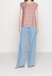 Ärmelloses Top mit Blumenmuster und Rüschenschultern, kombiniert mit hellblauen weit geschnittenen Jeans. Abgerundet mit schwarz-weißen Sandalen mit Riemchen.