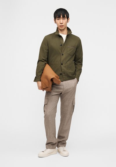 Άνδρας που φοράει ελιάς πράσινο σακάκι με κουμπιά, μπεζ παντελόνια καρό και λευκά αθλητικά παπούτσια, κρατώντας ένα διπλωμένο καφέ παλτό πάνω σε λευκό φόντο.