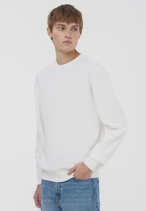 Maglione bianco a maniche lunghe con collo rotondo, realizzato in tessuto texturizzato, con polsini e orlo a coste, indossato con jeans in denim azzurri.
