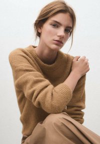 Suéter de lana en marrón claro con cuello redondo y puños acanalados, combinado con una falda beige mate, sobre un fondo neutro.