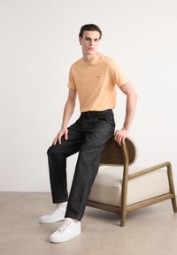 Orange crew-neck t-shirt med logo, parret med mørk denim jeans med synlige stikninger, og hvide sneakers på en træstol.