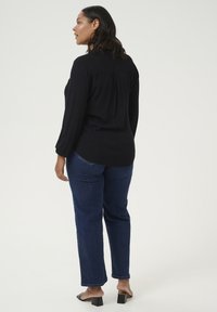 Blouse noire à manches longues avec un détail froncé dans le dos, associée à un jean droit en denim bleu et à une sandale à talons carrés noirs.