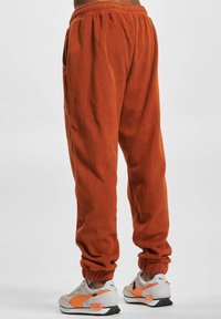 Orange fleece joggers with elastic waistband and cuffs. Features side pockets and a relaxed fit. Paired with white sneakers with orange accents.