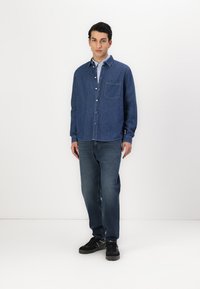 Donkerblauwe denim blouse met knopen, voorzien van een borstzak en een kraag, in combinatie met vervaagde blauwe jeans en zwarte sneakers.
