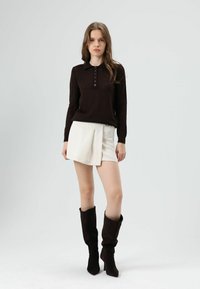 Donkerbruine gebreide trui met een polokraag en knoopsluiting, gecombineerd met een witte geplooide minirok en donkerbruine laarzen tot aan de knie.