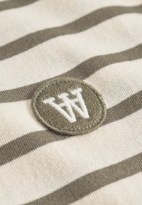 Material din bumbac cu dungi în nuanțe de crem și verde măsliniu, având un patch circular brodat cu logo-ul "W" în alb.