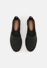 Paire d'espadrilles noires slip-on avec surpiqûres beige et petit logo Tommy Hilfiger sur le dessus et à l'intérieur de la semelle.
