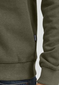 Sudadera verde oliva hecha de una tela suave. Puños y dobladillo acanalados con una pequeña etiqueta de marca en el lateral. Textura suave, ajuste casual.