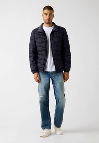 Navy puffer jacket with quilted design, zip closure, and gray collar, paired with light blue distressed jeans and white sneakers.