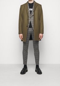 Olijfgroen jas over een grijs geruit pak met een zwart T-shirt. De schoenen zijn zwarte instappers. De outfit heeft een slim-fit broek en een stijlvolle uitstraling.