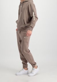 Sweat à capuche marron et joggings assortis en tissu doux. Le sweat à capuche comporte une poche avant et un logo ; les joggings ont des poignets élastiques et un logo. Baskets blanches.
